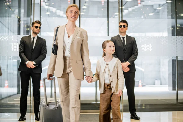 Personal security and protection, two bodyguards in suits and sunglasses standing near hotel entrance, happy mother and child holding hands and walking with luggage, entering lobby, luxury lifestyle — Stock Photo