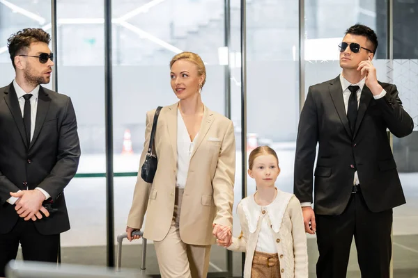 Personal security concept, two bodyguards in suits and sunglasses protecting happy blonde woman and child, luxury lifestyle, woman and girl holding hands and walking inside of hotel — Stock Photo