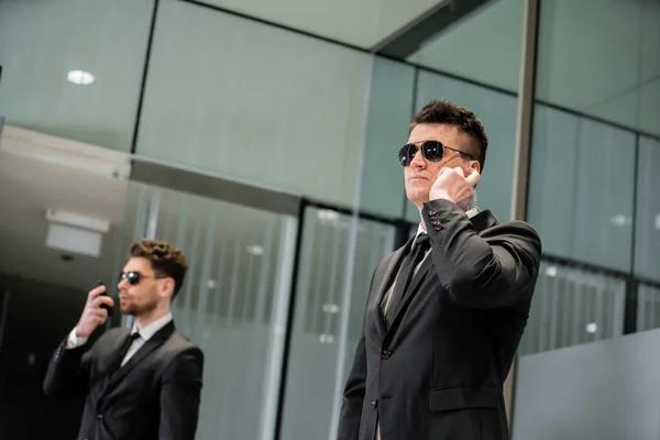 Hotel security concept, professional guards in suits and sunglasses standing in hotel lobby, handsome man with earpiece communicating with work partner, luxury hotel, vigilance, private bodyguards — Stock Photo