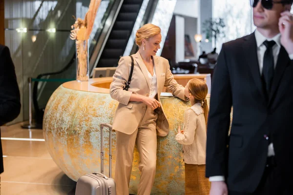 Personal security concept, blonde woman standing at reception desk with preteen daughter, rich lifestyle, family travel, bodyguard in suit and sunglasses standing on blurred foreground, safety — Stock Photo
