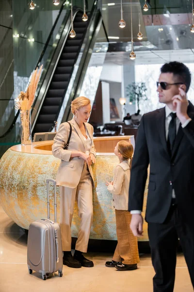 Personal safety concept, blonde woman standing at reception desk with preteen daughter, rich lifestyle, family travel, bodyguard in suit and sunglasses standing on blurred foreground, safety — Stock Photo