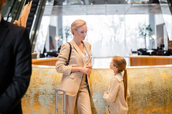 Familienreise, blonde Frau steht mit frühgeborener Tochter an der Rezeption, bespricht etwas beim Einchecken, Hotelbranche, Leibwächter im verschwommenen Vordergrund — Stockfoto