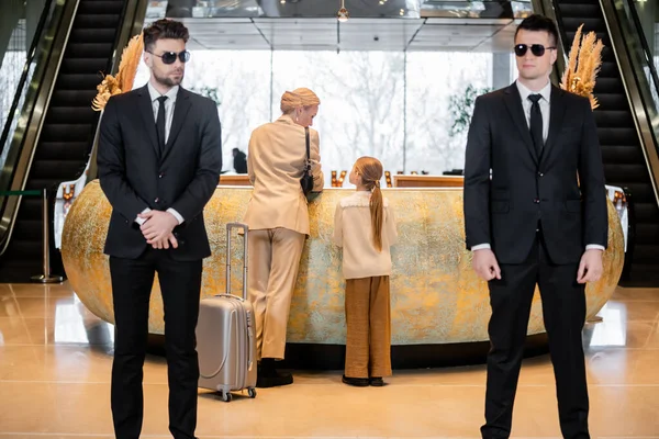 Personal security, luxury lifestyle, blonde mother with child standing at reception desk, woman and preteen girl during check in, bodyguards in suit and sunglasses protecting rich family in hotel — Stock Photo