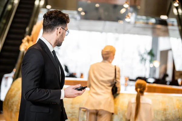 Bodyguard concept, handsome man in suit and tie holding radio transceiver, protecting clients on blurred background, looking at mother and daughter at reception desk, connection and safety — Stock Photo