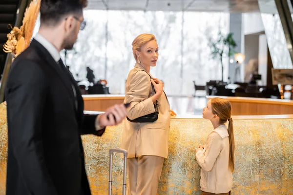 Personal security, lifestyle, blonde mother with child standing at reception desk, woman and preteen girl looking at camera during check in, bodyguard in suit standing on blurred foreground — Stock Photo