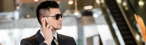 Bodyguard concept, handsome man in formal wear and tie touching earpiece in lobby of hotel, security, communication, hotel safety, career in security, profession, banner — Stock Photo