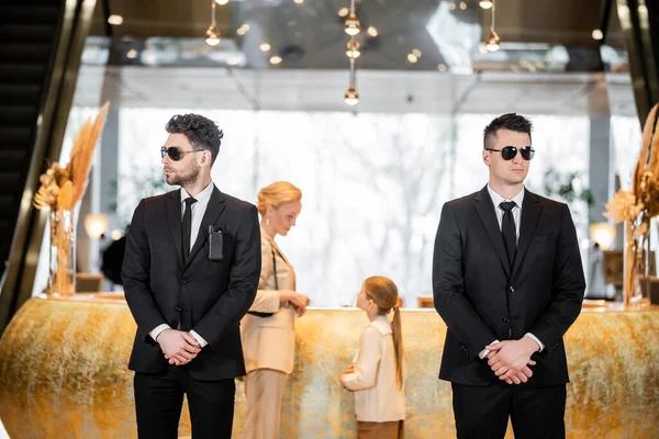 Personal security, handsome bodyguards in suits and sunglasses protecting mother and child in hotel lobby, luxury lifestyle, woman and girl standing at reception desk — Stock Photo