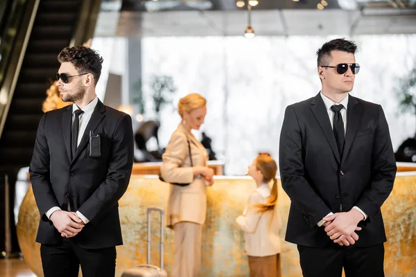 Personal security, handsome bodyguards in suits and sunglasses protecting life of mother and child in hotel lobby, rich and luxury lifestyle, woman and girl standing at reception desk — Stock Photo