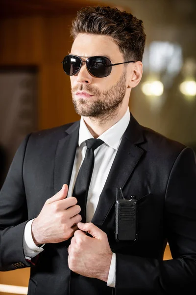 Handsome bodyguard, security guard in suit with tie and sunglasses standing in hotel, professional headshots, radio transceiver attached to jacket pocket, bearded man working in hotel security — Stock Photo