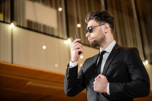 Good looking bodyguard, security worker in suit and sunglasses working in lobby of hotel, professional headshots, bearded man using radio transceiver while working in hotel — Stock Photo