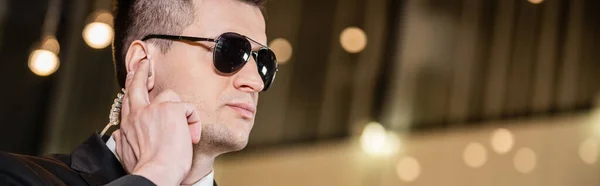 Handsome bodyguard in sunglasses, handsome man in suit and tie touching earpiece in lobby of hotel, security, career, communication, vigilance, private safety, hotel staff, banner — Stock Photo