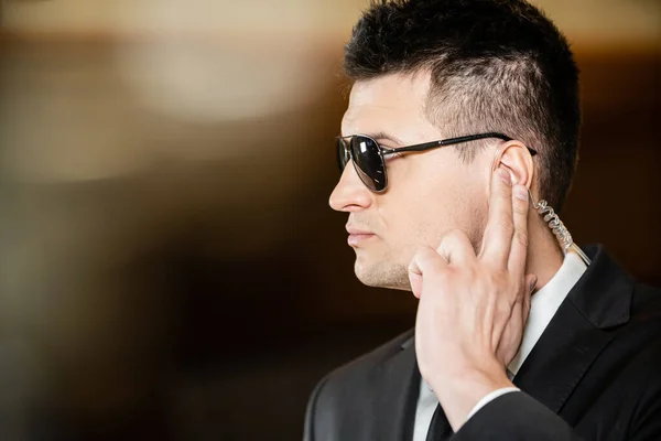 Handsome bodyguard in sunglasses, handsome man in suit and tie touching earpiece in lobby of hotel, security, career, communication, vigilance, private safety, hotel work — Stock Photo