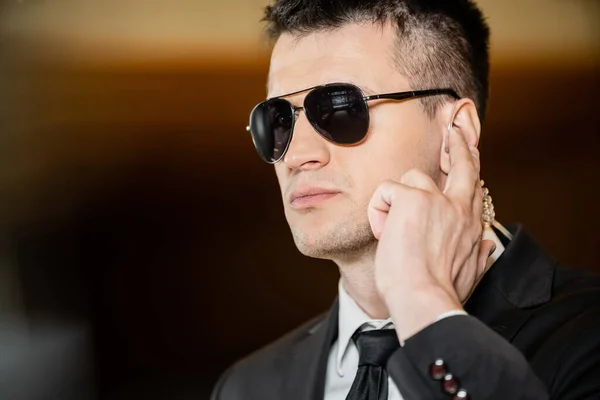 Handsome bodyguard in sunglasses, handsome man in suit and tie touching earpiece in lobby of hotel, security, career, communication, vigilance, private safety, hotel industry, male personnel — Stock Photo