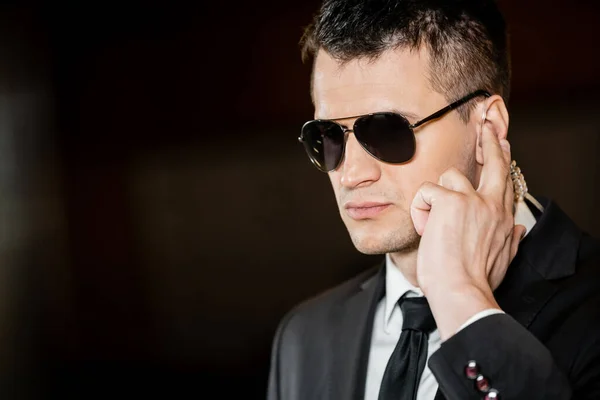 Good looking bodyguard in sunglasses, handsome man in suit touching earpiece in lobby of hotel, security, career, communication, vigilance, private safety, hotel security staff, male personnel — Stock Photo