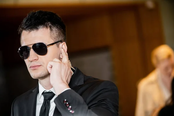 Handsome bodyguard in dark sunglasses, handsome man in suit and tie touching earpiece in lobby of hotel, security and career, communication, vigilance, private safety, hotel safety, male personnel — Stock Photo