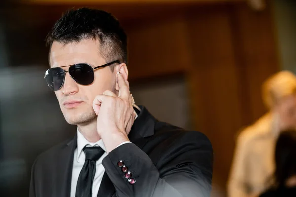 Bodyguard in dark sunglasses, handsome man in suit and tie touching earpiece in lobby of hotel, security, career, communication, vigilance, private safety, hotel safety, male personnel — Stock Photo