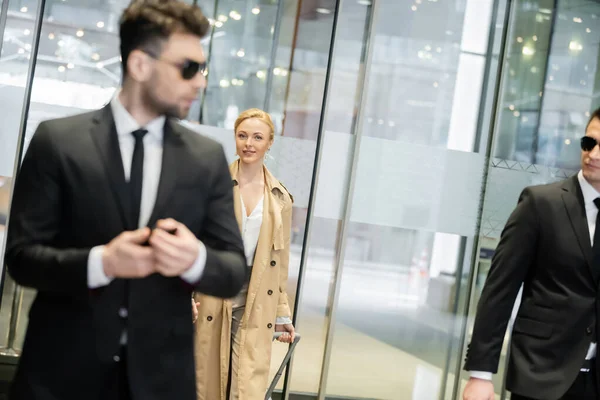 Seguridad privada, mujer rubia en gabardina que entra en el hotel, caminando con equipaje, dos guardaespaldas en ropa formal y gafas de sol en primer plano borroso, seguridad personal - foto de stock