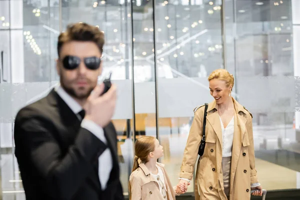 Bodyguard concept, luxury lifestyle, blonde woman and girl holding hands and walking inside of hotel, man in suit using radio transceiver, protecting clients on blurred foreground, personal security — Stock Photo