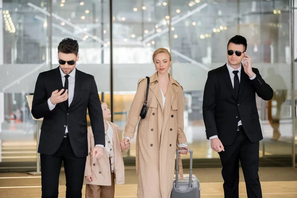 Personal security concept, two bodyguards in suits and sunglasses protecting blonde woman with child with luggage entering hotel, wearing autumnal clothes, luxury and rich lifestyle — Stock Photo