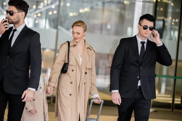 Privates Sicherheitskonzept, zwei Leibwächter in Anzügen und Sonnenbrillen schützen blonde Frau mit Kind mit Gepäck im Hotel, tragen herbstliche Kleidung, Luxus und reichen Lebensstil — Stockfoto