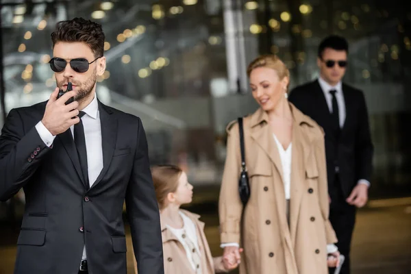 Handsome bodyguard communicating through walkie talkie, standing next to successful woman and preteen kid, private security, blonde mother and daughter in trench coats, safety and protection — Stock Photo