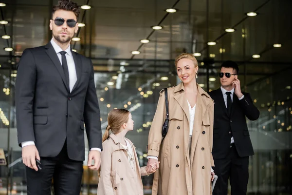 Two bodyguards protecting successful woman and kid, private security, happy blonde mother and daughter in trench coats near hotel, safety and protection, family travel, rich life — Stock Photo
