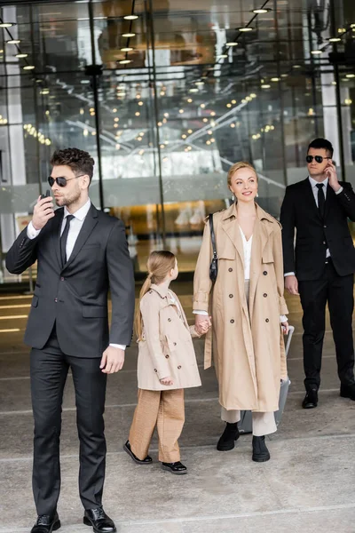 Handsome bodyguards protecting successful woman and preteen kid, private security, happy blonde mother and daughter in trench coats near hotel, safety and protection, family travel, rich life — Stock Photo