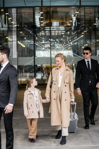 Professional bodyguards protecting successful woman and preteen kid, happy blonde mother and daughter in trench coats standing near hotel, safety and protection, private security — Stock Photo