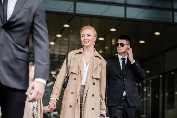 Professional bodyguards protecting smiling blonde woman and preteen kid, successful mother and daughter in trench coats standing near hotel, safety and protection, private security, guards — Stock Photo