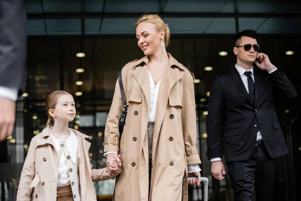 Professional bodyguards protecting happy woman and preteen kid, successful blonde mother and daughter in trench coats standing near hotel, safety and protection, private security — Stock Photo