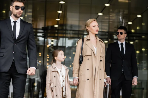Bodyguards walking next to blonde successful woman and preteen kid, entering hotel, private security, mother and daughter in trench coats, safety and protection, family travel, rich life — Stock Photo