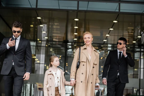 Bodyguards walking next to cheerful woman and preteen kid, entering hotel, private security, mother holding hands with daughter and wearing trench coats, safety and protection — Stock Photo