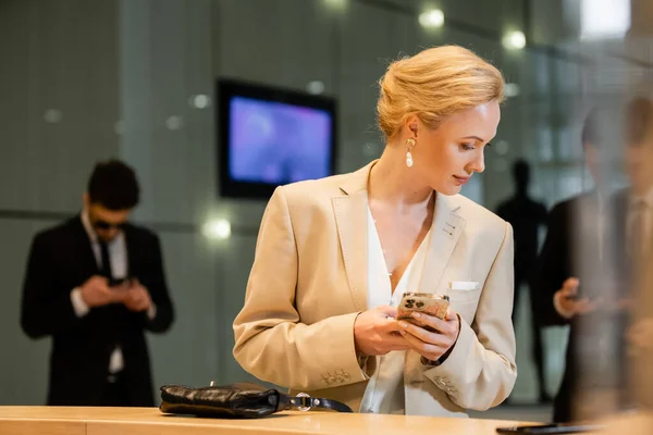 Personal security, blonde woman in suit holding smartphone near reception desk, two bodyguards standing on blurred background, digital age, hospitality industry, personal security — Stock Photo