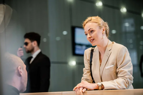 Polite businesswoman talking to receptionist in hotel, hospitality industry, blonde and beautiful woman communicating with hotel staff, private security, personal safety, bodyguard on background — Stock Photo