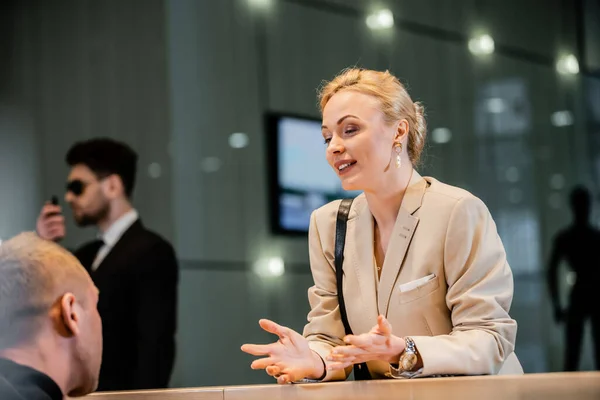 Polite businesswoman talking to receptionist in hotel, hospitality industry, blonde woman communicating with hotel personnel, private security, personal safety, bodyguard on background — Stock Photo