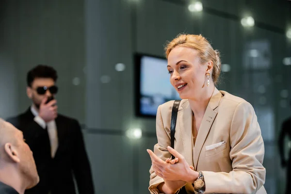 Polite businesswoman talking to receptionist in hotel, hospitality industry, blonde and charming woman communicating with hotel staff, private security, personal safety, bodyguard on background — Stock Photo