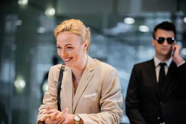 Happy blonde woman in formal wear standing at reception desk, personal security service concept, bodyguard in suit on blurred background, hotel industry, luxury travel, formal wear — Stock Photo