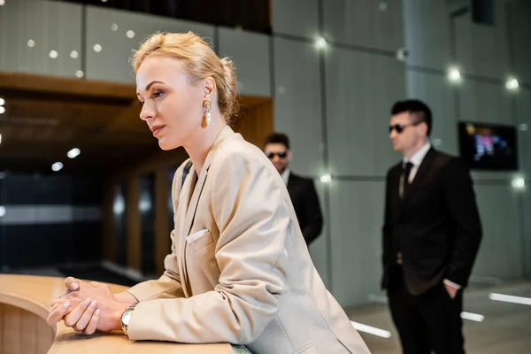 Successful blonde woman in formal wear standing at reception desk, personal security service concept, two bodyguards in suits on blurred background, hotel industry, luxury travel, formal wear — Stock Photo
