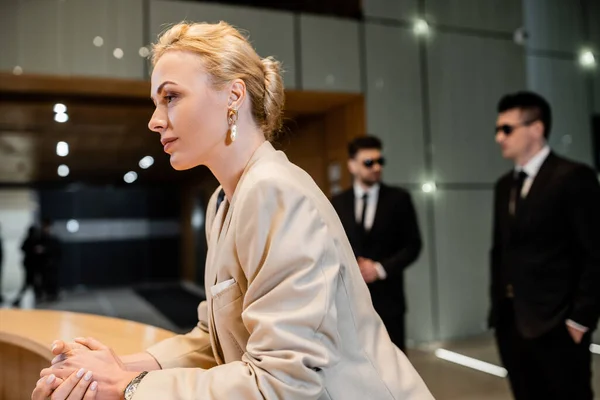 Blonde woman in suit standing at reception desk, personal security service concept, two bodyguards in suits standing on blurred background, hotel industry, luxury travel, formal wear — Stock Photo