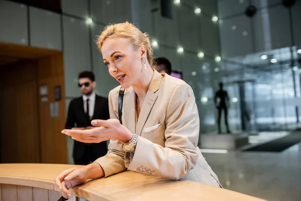 Blonde woman in suit gesturing while talking at reception desk, personal security service concept, bodyguards in suits standing on blurred background, hotel industry, safety during travel — Stock Photo