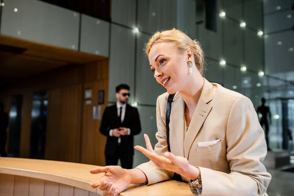 Polite blonde woman in suit gesturing while talking at reception desk, personal security service concept, bodyguard in suit standing on blurred background, hotel industry — Stock Photo