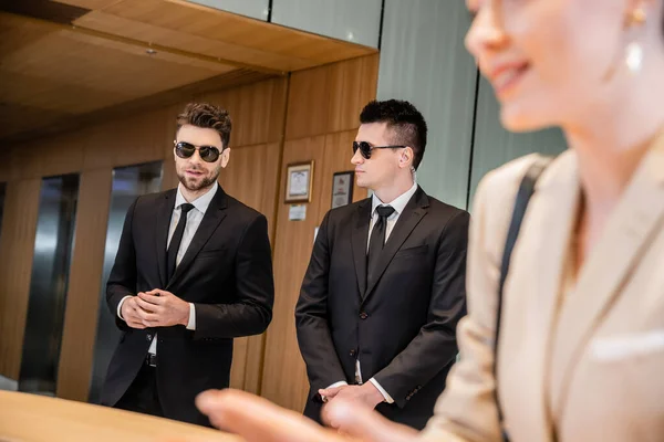 Handsome bodyguards protecting successful client in hotel, woman on blurred foreground, security service, personal safety, men in suits and sunglasses, hotel reception — Stock Photo