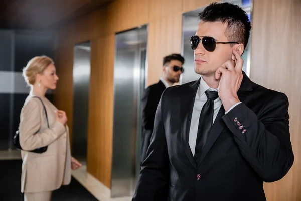 Bodyguard service, personal protection, blonde woman in formal wear standing near elevators, security personnel protecting successful businesswoman in hotel, handsome man with earpiece — Stock Photo