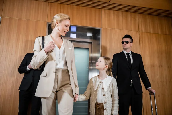 Happy mother and child holding hands while standing near hotel elevators and bodyguards in suits, personal protection, successful woman and preteen daughter, family travel, security service — Stock Photo