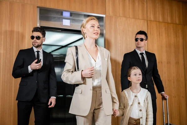 Blonde mother and child holding hands while standing near hotel elevator and bodyguards in suits, personal protection, successful woman and preteen daughter, family travel, security service — Stock Photo