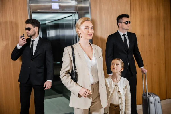 Successful mother and child holding hands while standing near hotel elevator and bodyguards in suits, personal protection, woman and preteen daughter, family travel, security service — Stock Photo