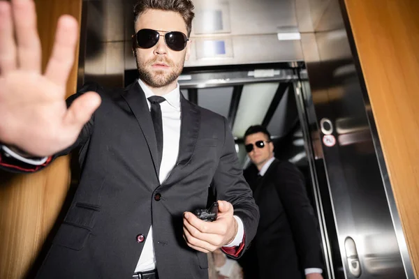 Private security concept, handsome bodyguard in sunglasses and suit showing stop, no gesture to camera, protecting private space, hotel lobby, standing next to work partner in elevator — Stock Photo