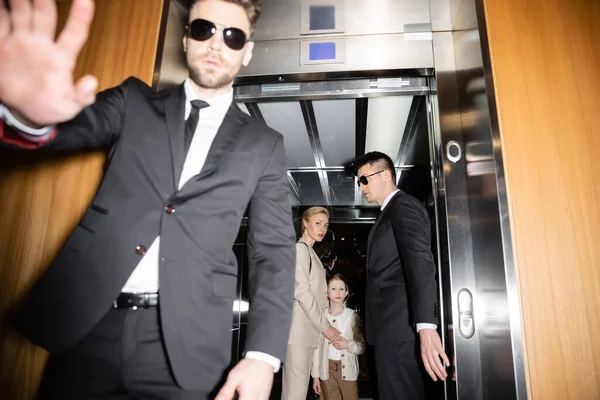 Private security concept, handsome bodyguard in sunglasses and suit showing stop, no gesture to camera, protecting private space of famous clients in elevator of hotel, blurred mother and daughter — Stock Photo