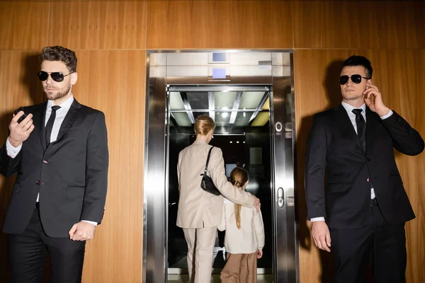 Seguridad privada, madre e hija entrando en ascensor de lujoso hotel, dos guardaespaldas protegiendo su seguridad, hombres guapos en trajes y gafas de sol trabajando en servicio de seguridad personal - foto de stock