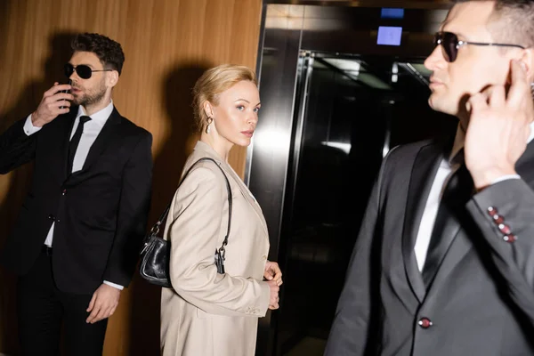 Persönliches Sicherheits- und Schutzkonzept, blonde und erfolgreiche Frau mit Handtasche, die neben Leibwächtern in Anzügen und Sonnenbrillen im Fahrstuhl steht, Luxushotel — Stockfoto